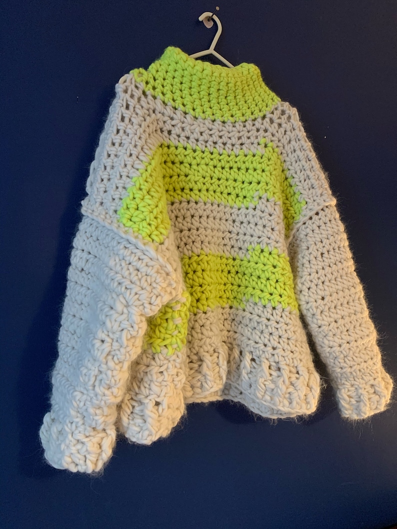 May include: A chunky crochet sweater featuring a high neckline and long sleeves. The garment is crafted from thick yarn in alternating horizontal stripes of off-white and vibrant neon yellow, showcasing a textured, hand-stitched knit pattern.