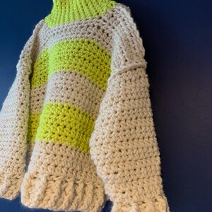 May include: A chunky crochet sweater featuring a high neck and long sleeves, crafted with thick yarn in alternating horizontal stripes of neon yellow and off-white. The garment has a textured, hand-stitched appearance with ribbed cuffs and hem.