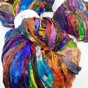 Könnte beinhalten: Mehrere Bündel farbenfroher Seidenwolle. Die Wolle weist eine Mischung aus Blau, Lila, Orange und Grün auf. Jedes Bündel ist mit einem weißen Etikett mit der Aufschrift "Yarn Yarn" versehen. Die Wolle hat ein strukturiertes, ausgefranstes Aussehen.