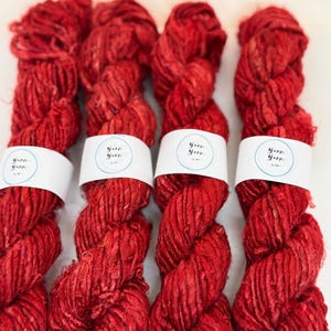 Könnte beinhalten: Vier Stränge leuchtend roter Wolle werden gezeigt, jeder eng gewickelt und mit einem weißen Band mit dem Text "Yarn Yarn Yarn" versehen. Die Wolle hat ein strukturiertes Aussehen, das auf eine Naturfaser hindeutet, und ist vor einem weißen Hintergrund angeordnet.