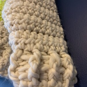 May include: A chunky hand-knit textile featuring thick, off-white yarn worked in a prominent ribbed stitch pattern. A small section of bright neon yellow yarn is visible along the upper left edge, contrasting with the neutral cream-colored fibers.