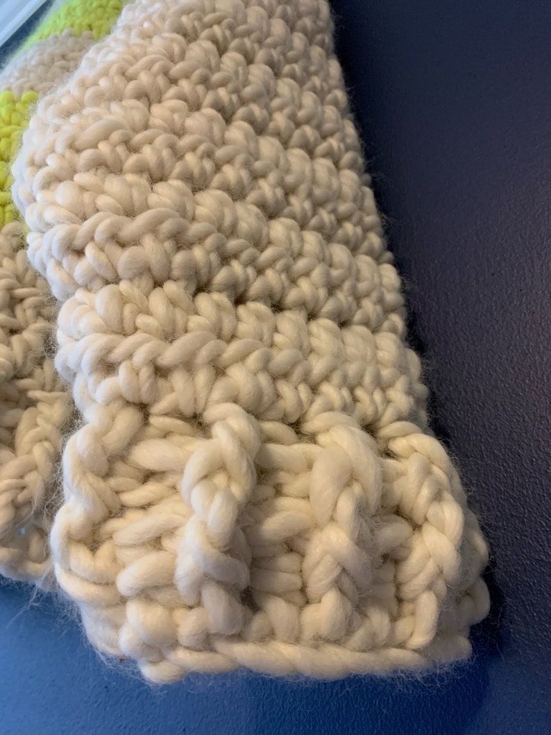 May include: A chunky hand-knit textile featuring thick, off-white yarn worked in a prominent ribbed stitch pattern. A small section of bright neon yellow yarn is visible along the upper left edge, contrasting with the neutral cream-colored fibers.