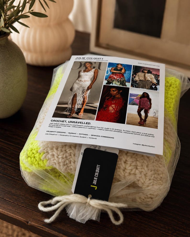 May include: A folded neon yellow and cream crochet textile packaged in clear plastic. A white informational card titled JULIE COLQUITT CROCHET, UNRAVELLED is attached, featuring five photographs of crochet garments and a black branded hang tag tied with string.