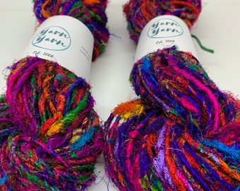 May include: Four colorful yarn skeins with vibrant hues of purple, orange, green, and blue. Each skein has a white label with the text "yarn yarn" and "Est. 2008". The yarn appears to be a textured, multi-colored fiber.