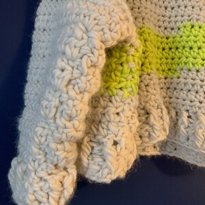 May include: A chunky crochet sweater featuring a thick, off-white yarn base with horizontal stripes in a vibrant neon yellow. The garment is constructed with a prominent, textured stitch pattern that creates a dense, soft, and layered appearance.