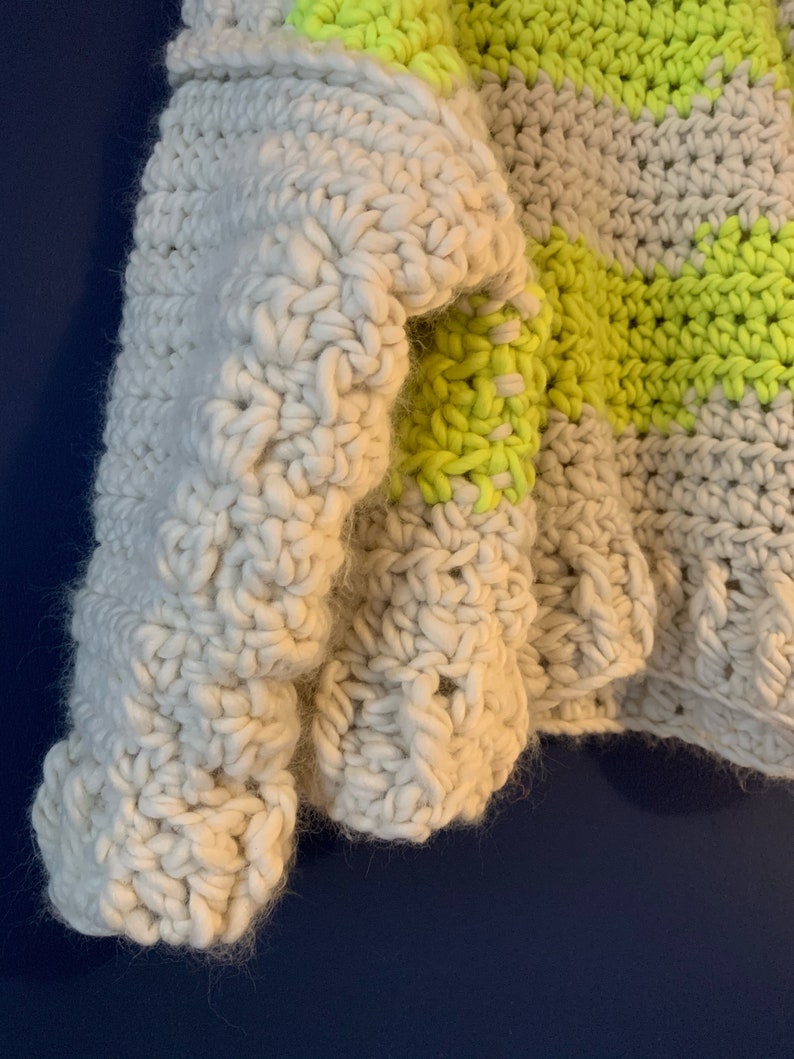 May include: A chunky crochet sweater featuring a thick, off-white yarn base with horizontal stripes in a vibrant neon yellow. The garment is constructed with a prominent, textured stitch pattern that creates a dense, soft, and layered appearance.