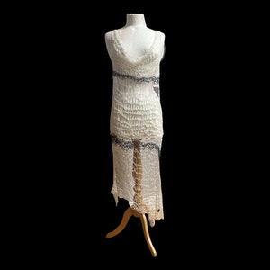 Peut inclure: Une robe sans manches arrivant au genou, confectionn&eacute;e dans un tissu crochet&eacute; ajour&eacute; blanc cass&eacute;. Le v&ecirc;tement pr&eacute;sente un d&eacute;collet&eacute; drap&eacute; et deux bandes horizontales de d&eacute;tails complexes gris fonc&eacute; rappelant la dentelle sur la poitrine.