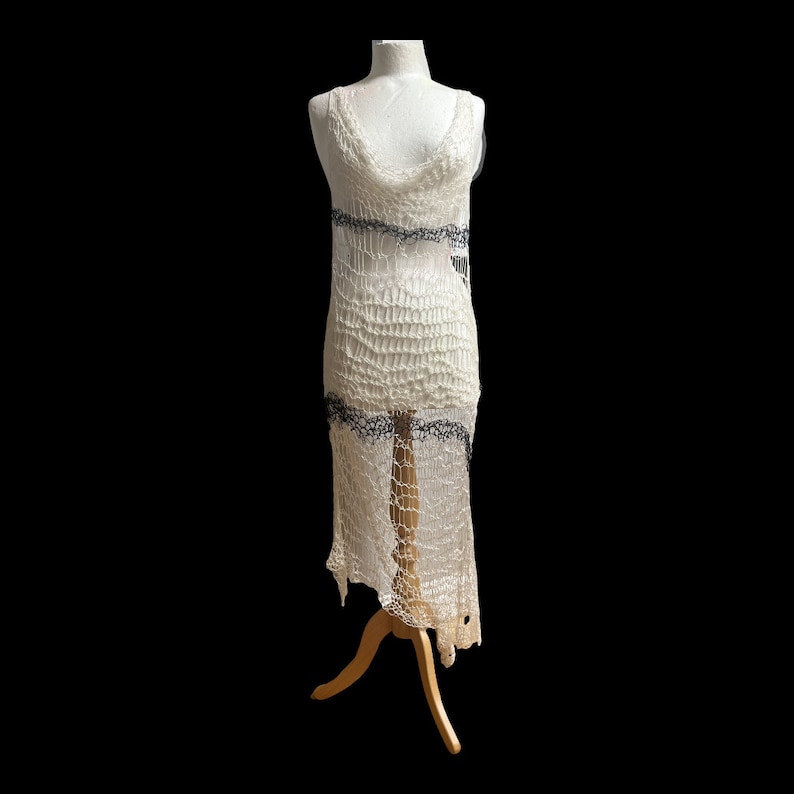 Peut inclure: Une robe sans manches arrivant au genou, confectionn&eacute;e dans un tissu crochet&eacute; ajour&eacute; blanc cass&eacute;. Le v&ecirc;tement pr&eacute;sente un d&eacute;collet&eacute; drap&eacute; et deux bandes horizontales de d&eacute;tails complexes gris fonc&eacute; rappelant la dentelle sur la poitrine.