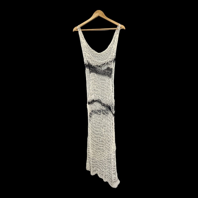 Peut inclure: Une robe longue sans manches en tricot ajour&eacute; blanc avec une texture l&acirc;che semblable &agrave; un filet. Le v&ecirc;tement pr&eacute;sente des accents de fil noir ondul&eacute;s et horizontaux sur la poitrine et la taille, suspendu &agrave; un cintre en bois.