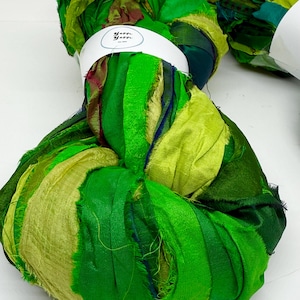 May include: Several bundles of green silk ribbon yarn in various shades, from light lime to deep forest green. The yarn is wrapped with a white band that has the text "Yarn Yarn" printed on it. The texture of the yarn appears soft and slightly frayed.