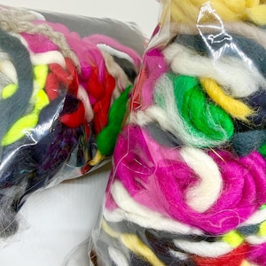Pode incluir: Duas sacolas plásticas transparentes cheias de lã colorida. A lã inclui tons de rosa, branco, verde, amarelo, vermelho e cinza. As sacolas são parcialmente visíveis, mostrando a textura e variedade da fibra.
