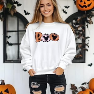 May include: White sweatshirt with the word "Boo" in orange letters and two pumpkin designs. The pumpkins have Mickey Mouse ears. The model is wearing black jeans and is standing in front of a white door with a pumpkin and Halloween decorations.