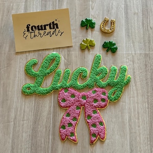 May include: A St. Patrick's Day themed arrangement featuring a green "Lucky" sign with a pink polka-dotted bow, shamrocks, a gold horseshoe, and a card with the text "fourth & threads".