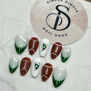 May include: A set of ten press-on nails featuring a football theme. The design includes brown glitter footballs with white laces, white nails with green mascot graphics, and white nails with green French tips. A circular logo reads Simple Dimple Nail Shop.