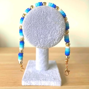 May include: A beaded bracelet with a mix of blue, white, and gold beads. The bracelet is displayed on a white stand. The beads are flat and disc-shaped, with a gold clasp.