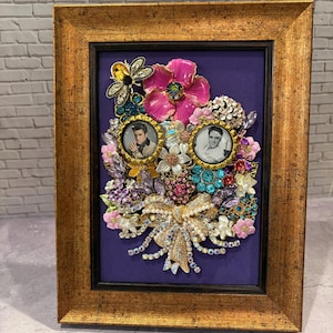 May include: Framed artwork featuring two small portraits surrounded by a vibrant arrangement of brooches and jewels. The frame is gold-toned, and the background is a deep purple. The arrangement includes pink, blue, and clear gemstones.