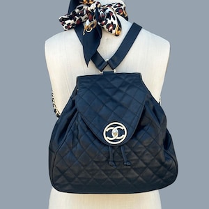 May include: Black quilted leather backpack with a gold-tone logo on the front flap. The backpack has a drawstring closure and chain straps. A patterned scarf is draped over the mannequin's neck.