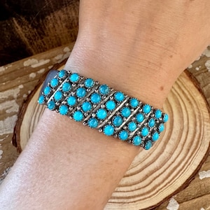 May include: A silver cuff bracelet featuring rows of vibrant turquoise stones. The bracelet is set against a wooden background, showcasing the bright blue of the stones. The bracelet is approximately 1 inch wide.