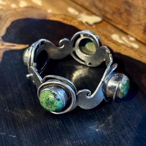Op de afbeelding: Een zilveren armband met een reeks ronde zettingen, elk voorzien van een ronde, gevlekte groene steen. Het metalen frame heeft een gladde, gepolijste afwerking met gebogen, golfachtige uitsparingen die de steenzettingen in een lus verbinden.