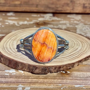May include: A silver cuff bracelet featuring a large, oval-shaped spiny oyster shell cabochon with vibrant orange and white striations. The metal band consists of three parallel silver wires that meet at decorative, fan-shaped silver accents.