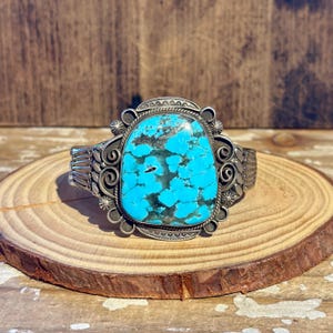May include: A silver cuff bracelet featuring a large, irregular, bright blue turquoise stone with dark matrix patterns. The metal setting is decorated with intricate scrollwork, twisted wire details, and small stamped sunburst accents.
