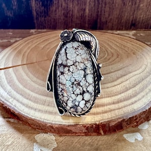 May include: A silver-toned ring featuring an oval, patterned stone with a mix of white, brown, and blue-green hues. The ring has a floral accent and leaf-like details, showcasing a detailed, nature-inspired design. The stone is approximately 1.5 inches long.