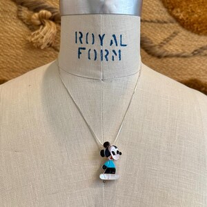 K&ouml;nnte beinhalten: Eine silberne Halskette mit einem Anh&auml;nger, der eine Zeichentrickfigur in Schwarz, Wei&szlig;, Blau und Rosa zeigt. Der Anh&auml;nger h&auml;ngt an einer Silberkette, pr&auml;sentiert auf einer Schaufensterpuppe. Der Text "ROYAL FORM" ist sichtbar.