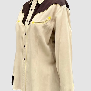 May include: A long-sleeved button-down shirt in beige with dark brown shoulder yokes and cuffs. The shirt features a pointed collar, a front button placket, and two chest pockets, all accented with thin, bright yellow piping along the seams.