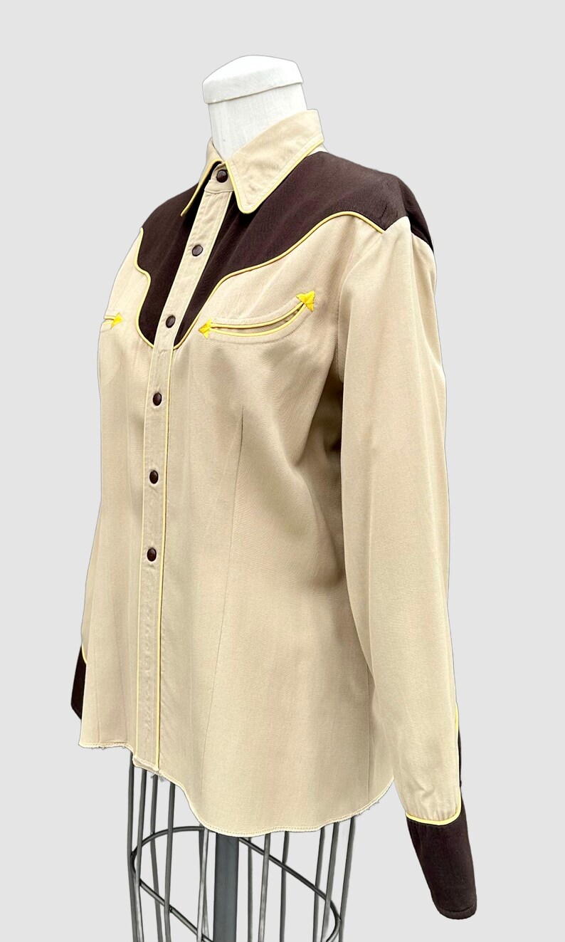 May include: A long-sleeved button-down shirt in beige with dark brown shoulder yokes and cuffs. The shirt features a pointed collar, a front button placket, and two chest pockets, all accented with thin, bright yellow piping along the seams.