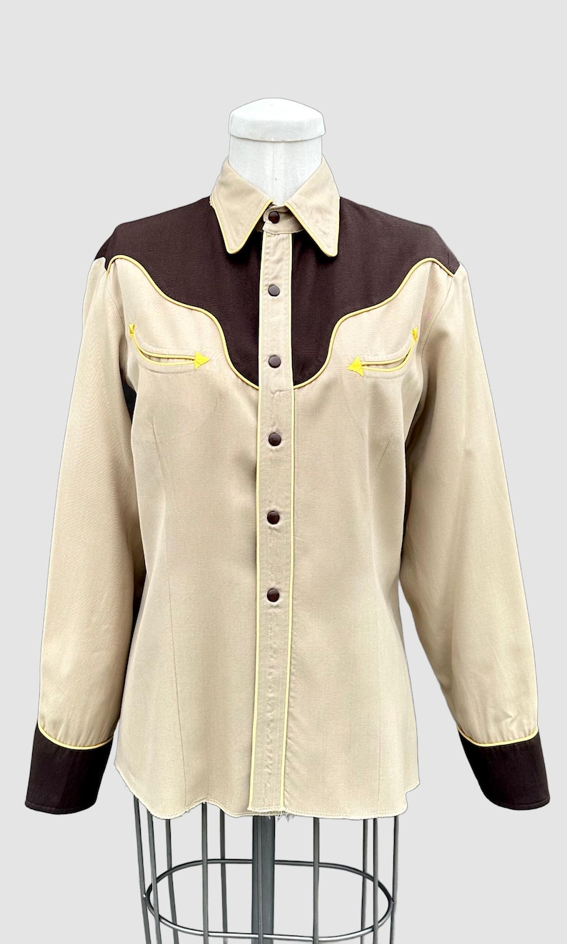 May include: A long-sleeve button-down shirt featuring a tan body with dark brown yokes and cuffs. The shirt includes yellow piping accents along the yokes and pockets, a pointed collar, and dark brown buttons down the front placket.