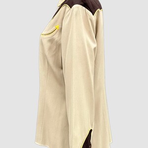 May include: A long-sleeved button-up shirt in beige with dark brown shoulder yokes and cuffs. The shirt features bright yellow piping along the yokes, cuffs, and a single chest pocket. It has a pointed collar and dark brown buttons on the cuffs.