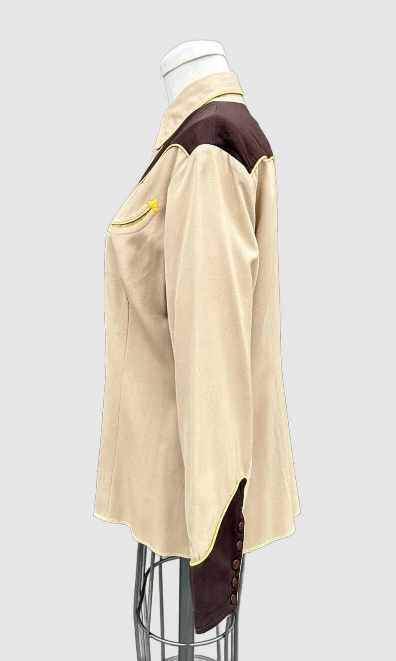 May include: A long-sleeved button-up shirt in beige with dark brown shoulder yokes and cuffs. The shirt features bright yellow piping along the yokes, cuffs, and a single chest pocket. It has a pointed collar and dark brown buttons on the cuffs.