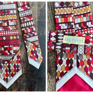 May include: A necktie featuring a geometric pattern of circles, diamonds, and rectangles in shades of red, orange, brown, and white. A yellow label on the back reads FLORENCE-MADE IN ITALY EMILIO PUCCI 100% PURE SILK. The tie has a pointed end.