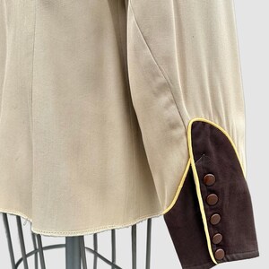 May include: A beige long-sleeved jacket featuring a dark brown cuff with yellow piping trim. The cuff is detailed with five small, round, dark brown buttons arranged in a vertical line along the outer edge of the sleeve opening.