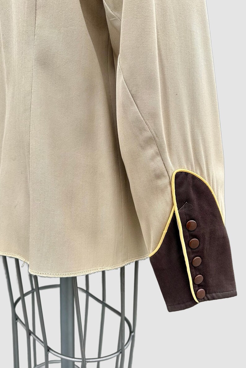 May include: A beige long-sleeved jacket featuring a dark brown cuff with yellow piping trim. The cuff is detailed with five small, round, dark brown buttons arranged in a vertical line along the outer edge of the sleeve opening.