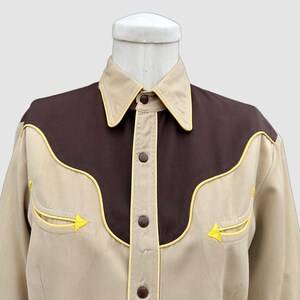 May include: A long-sleeved button-down shirt featuring a tan base colour with a dark brown yoke across the chest and shoulders. The shirt is accented with bright yellow piping along the yoke, collar, and two chest pockets, and has brown buttons.