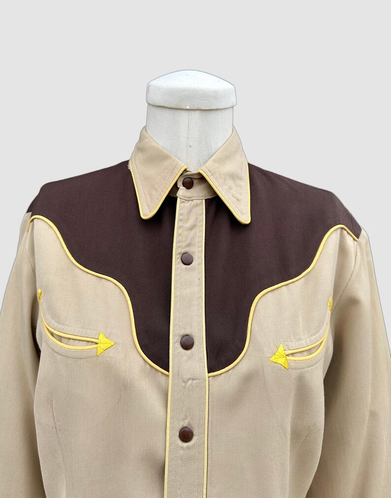 May include: A long-sleeved button-down shirt featuring a tan base colour with a dark brown yoke across the chest and shoulders. The shirt is accented with bright yellow piping along the yoke, collar, and two chest pockets, and has brown buttons.