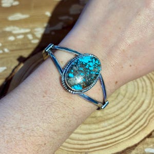 May include: A silver cuff bracelet featuring a central oval turquoise stone with dark matrix patterns. The metal band has a split design and a textured border surrounding the stone, creating a detailed frame for the vibrant blue and brown gemstone.