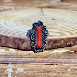 May include: A silver ring with a rectangular, vibrant red coral center stone. The ring features an ornate, swirling design around the coral and on the band, set against a natural wood background.