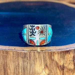 Può includere: Anello in argento con intarsi turchesi, corallo e neri. L'anello presenta disegni geometrici, tra cui un sole, una figura e altre forme. L'anello ha una fascia larga e una finitura lucida.