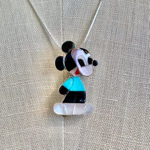 K&ouml;nnte beinhalten: Eine Halskette mit einem Mickey Mouse-Anh&auml;nger. Der Anh&auml;nger zeigt ein stilisiertes Mickey Mouse-Design mit schwarzen, wei&szlig;en, roten und t&uuml;rkisfarbenen Akzenten. Der Anh&auml;nger h&auml;ngt an einer d&uuml;nnen Silberkette.