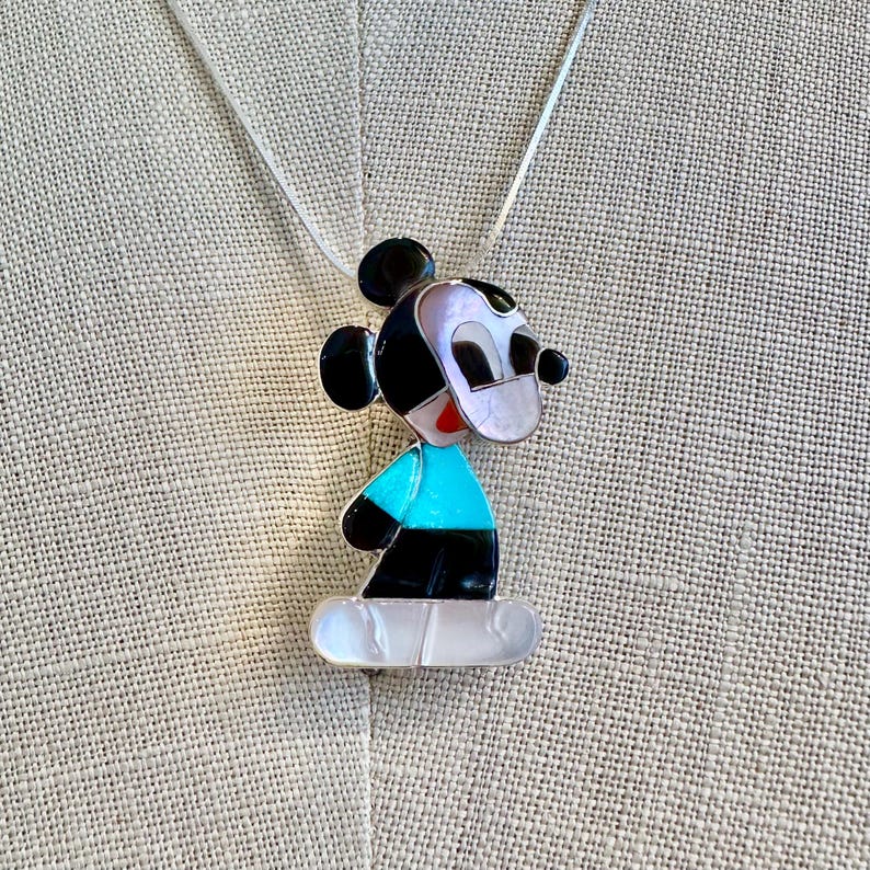 K&ouml;nnte beinhalten: Eine Halskette mit einem Mickey Mouse-Anh&auml;nger. Der Anh&auml;nger zeigt ein stilisiertes Mickey Mouse-Design mit schwarzen, wei&szlig;en, roten und t&uuml;rkisfarbenen Akzenten. Der Anh&auml;nger h&auml;ngt an einer d&uuml;nnen Silberkette.