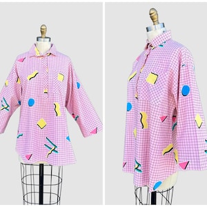 Puede incluir: Una camisa de manga larga con botones que presenta un patrón de cuadrícula rosa sobre un fondo rosa claro. La camisa está decorada con formas geométricas dispersas en amarillo, azul y rosa, incluyendo círculos, cuadrados y líneas onduladas.