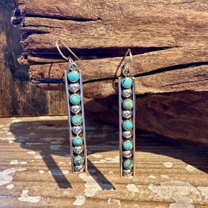 May include: A pair of silver-toned dangle earrings. Each earring features a rectangular design with a row of turquoise-colored stones and silver beads. The earrings are displayed against a rustic wooden background.