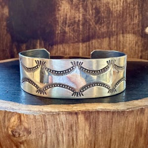 May include: A silver cuff bracelet with an intricate stamped design. The bracelet features a series of curved patterns with dotted and linear details. The metal has a polished finish, reflecting the light. The bracelet is displayed on a wooden surface.
