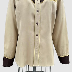 May include: A long-sleeved button-down shirt in beige with dark brown yokes and cuffs. The shirt features yellow piping accents along the yokes, cuffs, and front placket. It has dark brown buttons down the centre and two decorative chest pockets.