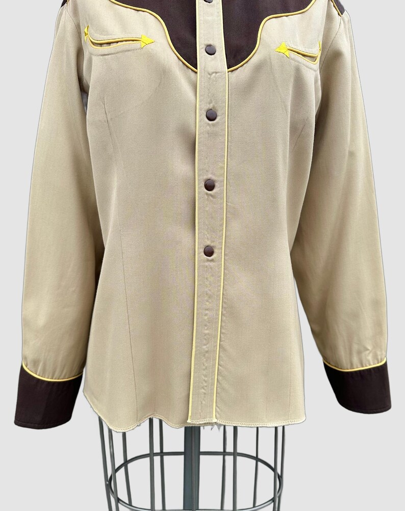 May include: A long-sleeved button-down shirt in beige with dark brown yokes and cuffs. The shirt features yellow piping accents along the yokes, cuffs, and front placket. It has dark brown buttons down the centre and two decorative chest pockets.