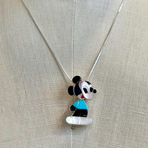 K&ouml;nnte beinhalten: Eine silberne Halskette mit einem Mickey Mouse Anh&auml;nger. Der Anh&auml;nger ist schwarz, wei&szlig; und blau, mit einer Basis aus Perlmutt. Die Halskette wird auf einer Schaufensterpuppe gezeigt.