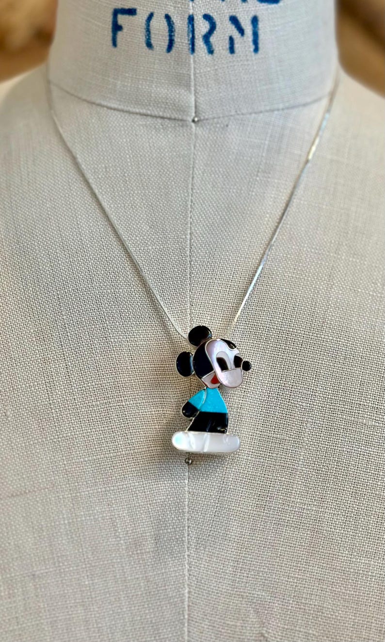 K&ouml;nnte beinhalten: Eine silberne Halskette mit einem Mickey Mouse Anh&auml;nger. Der Anh&auml;nger ist schwarz, wei&szlig; und blau, mit einer Basis aus Perlmutt. Die Halskette wird auf einer Schaufensterpuppe gezeigt.