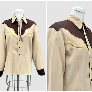 May include: A long-sleeved button-down shirt featuring a tan body with dark brown shoulder yokes and cuffs. The shirt includes yellow piping accents along the yokes and pockets, a pointed collar, and a bolo tie with a central decorative stone.